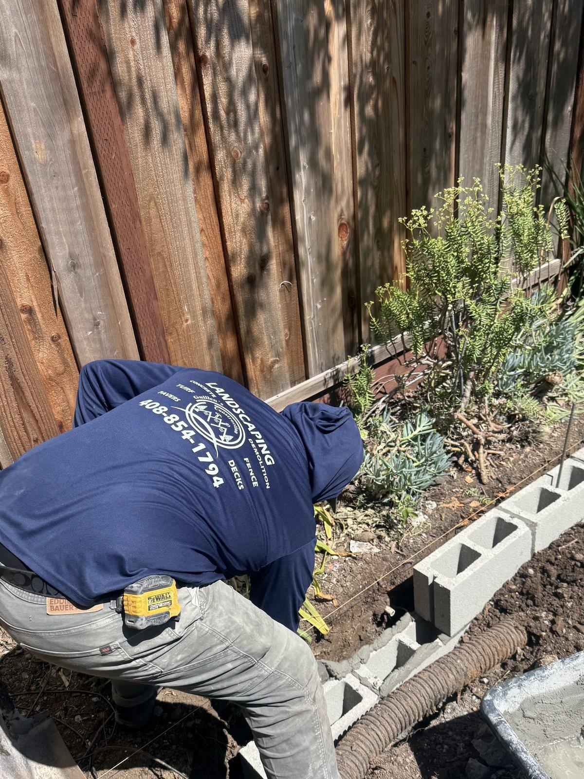 A Guillen's Construction crew member in a branded work shirt, setting concrete blocks by hand for a retaining wall along a fence line
