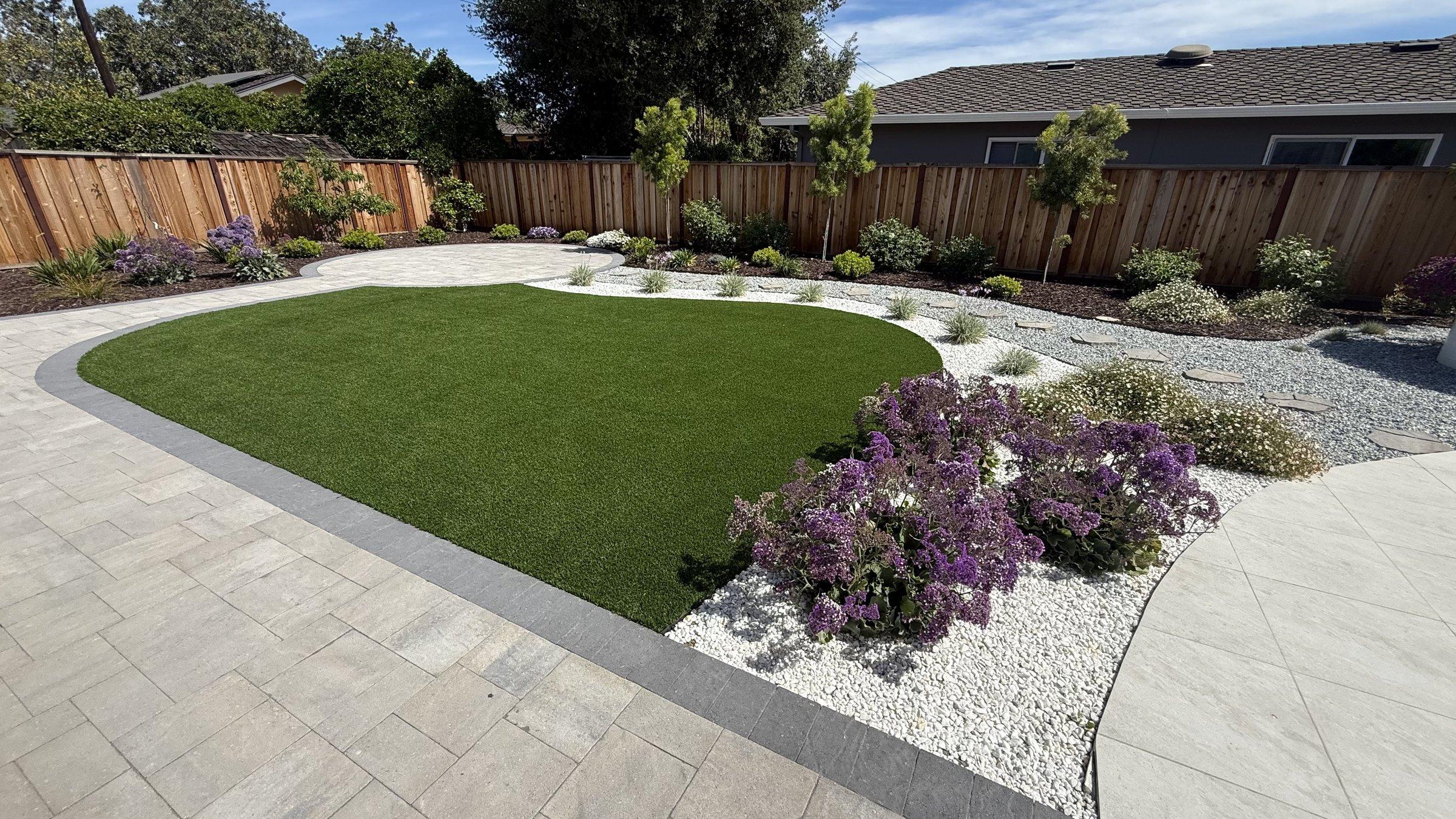 A backyard finished by Guillen's Construction — a curved paver patio wrapping an oval artificial turf lawn, with flowering planting beds against a redwood fence