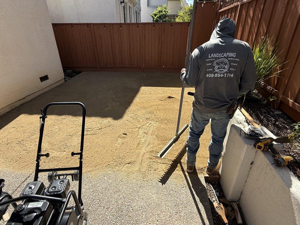 A crew member in a Guillen's Landscaping work hoodie spreading and screeding base material across a fenced side yard