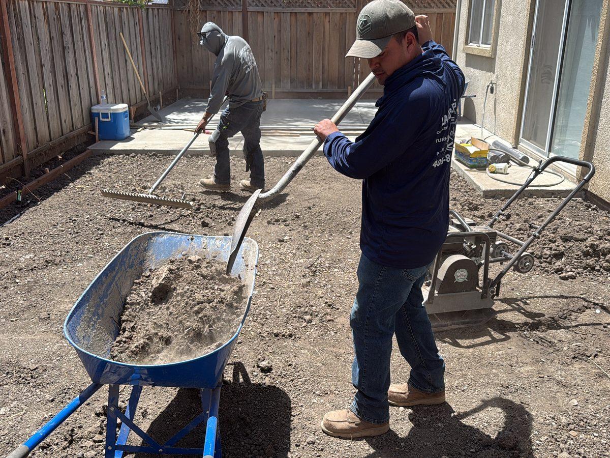 Two Guillen's Construction crew members grading a backyard, one shovelling from a wheelbarrow while the other rakes the base level