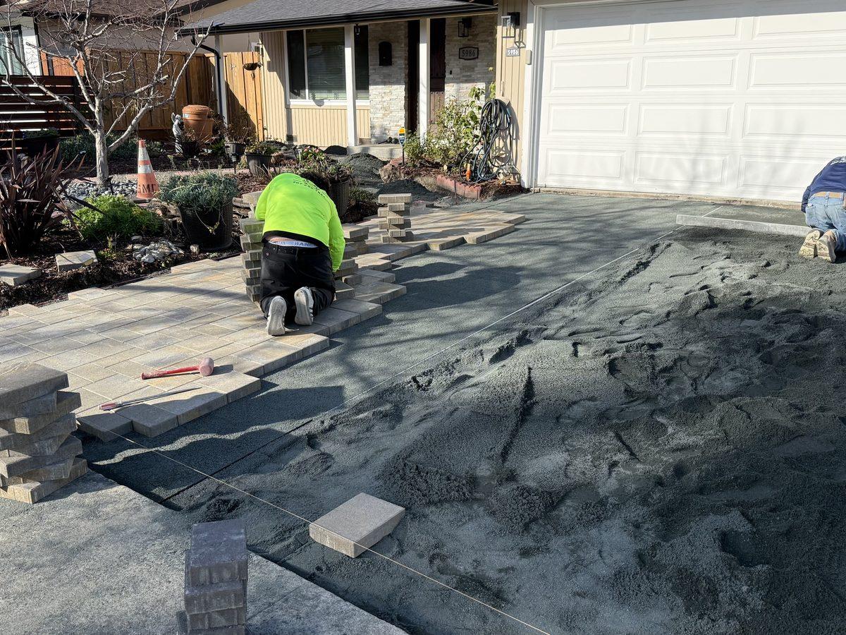 Two crew members kneeling on a driveway, setting interlocking pavers into the sand bed by hand