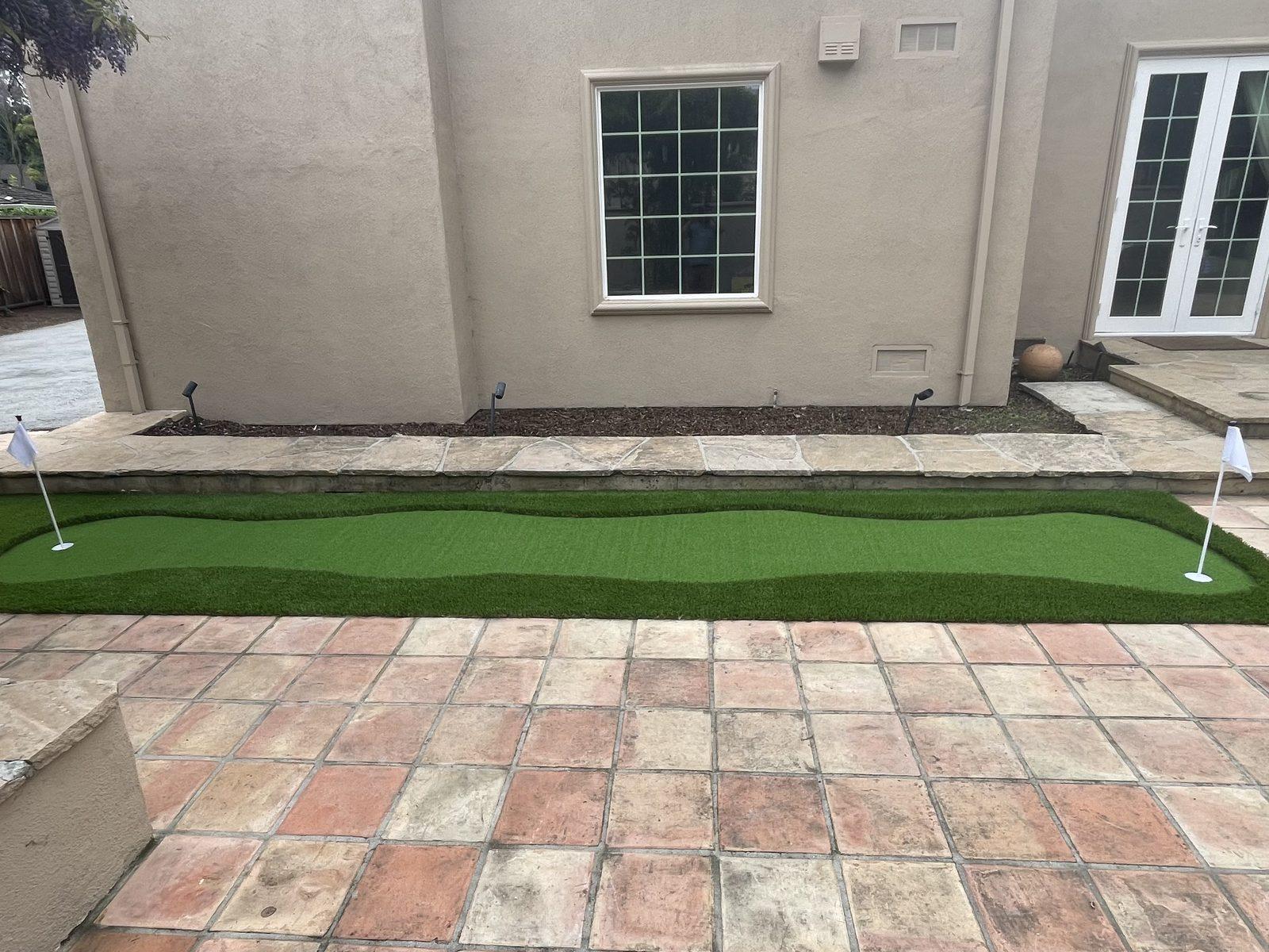 Finished backyard putting green with a flag pin, laid alongside a tiled patio