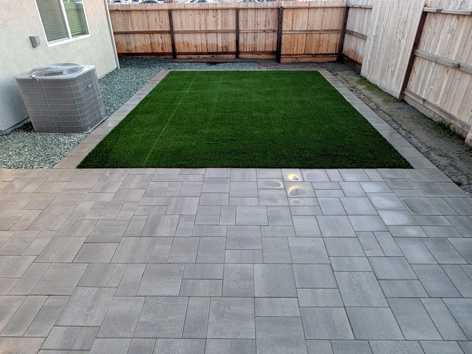 Rectangular artificial turf lawn set into a full paver patio, framed by a wood fence