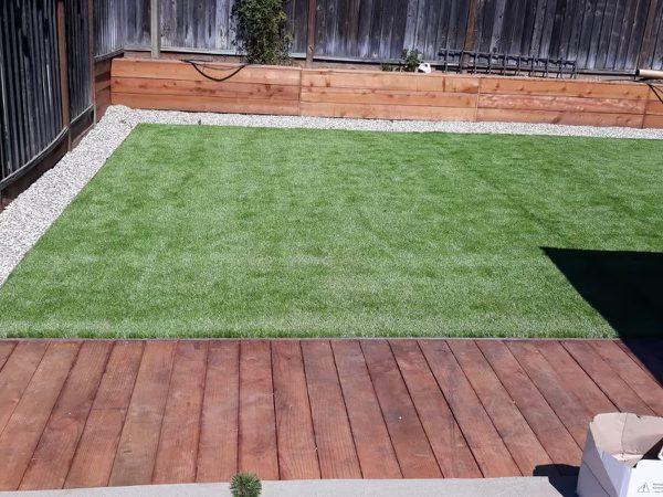 Backyard artificial turf lawn bordered by pale gravel, with a freshly built redwood deck along one edge